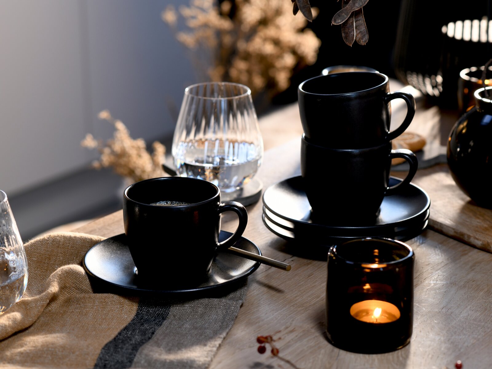 Schwarzes Kaffeetassen- und Untertassen-Set auf rustikalem Holztisch, eine Tasse im Vordergrund und zwei gestapelte Tassen rechts, seitliche Perspektive mit warmem Kerzenlicht und Glas im Hintergrund.