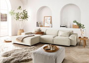 Helle, moderne Wohnzimmer-Ecke mit einer großen, beigen Eckcouch in L-Form, dekoriert mit Kissen und einer Decke. Im Vordergrund ein passender Hocker mit Tablett und Tassen. Die Perspektive zeigt die Couch von vorne, mit einem großen Fenster und Pflanzen