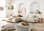 Helle, moderne Wohnzimmer-Ecke mit einer großen, beigen Eckcouch in L-Form, dekoriert mit Kissen und einer Decke. Im Vordergrund ein passender Hocker mit Tablett und Tassen. Die Perspektive zeigt die Couch von vorne, mit einem großen Fenster und Pflanzen 