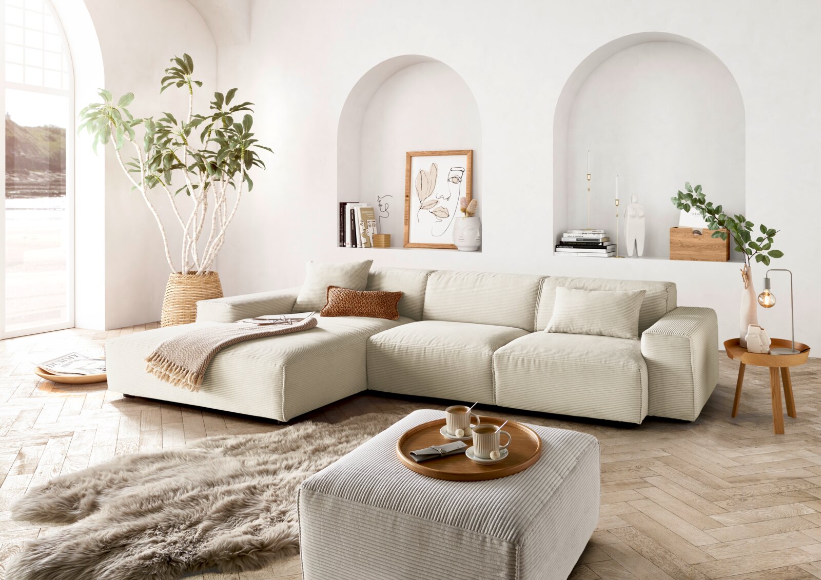Helle, moderne Wohnzimmer-Ecke mit einer großen, beigen Eckcouch in L-Form, dekoriert mit Kissen und einer Decke. Im Vordergrund ein passender Hocker mit Tablett und Tassen. Die Perspektive zeigt die Couch von vorne, mit einem großen Fenster und Pflanzen 