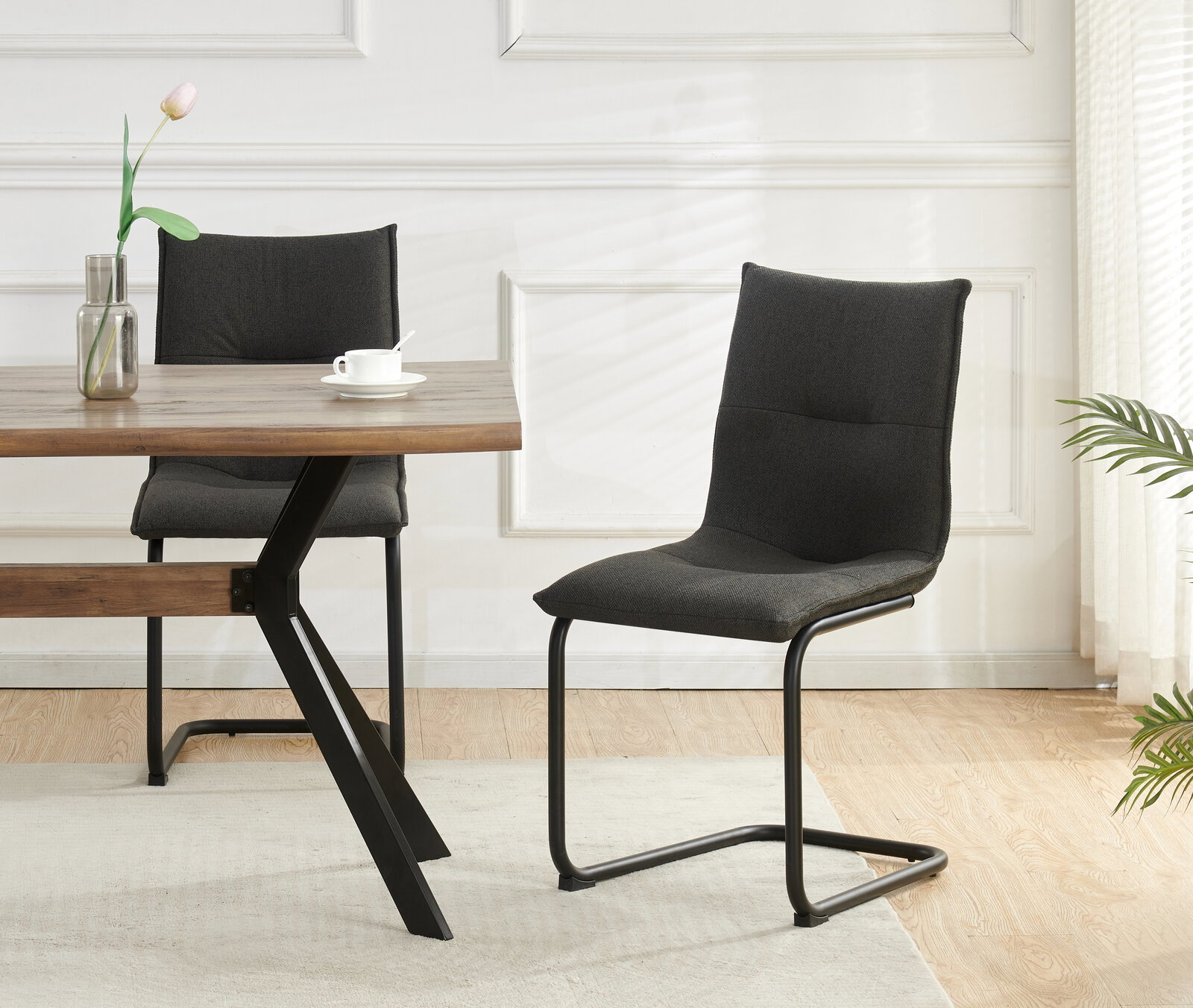 Moderner Esszimmerstuhl mit dunkelgrauem Stoffbezug und schwarzem Freischwinger-Metallgestell, seitliche Frontansicht im hellen Wohnraum neben Holztisch.