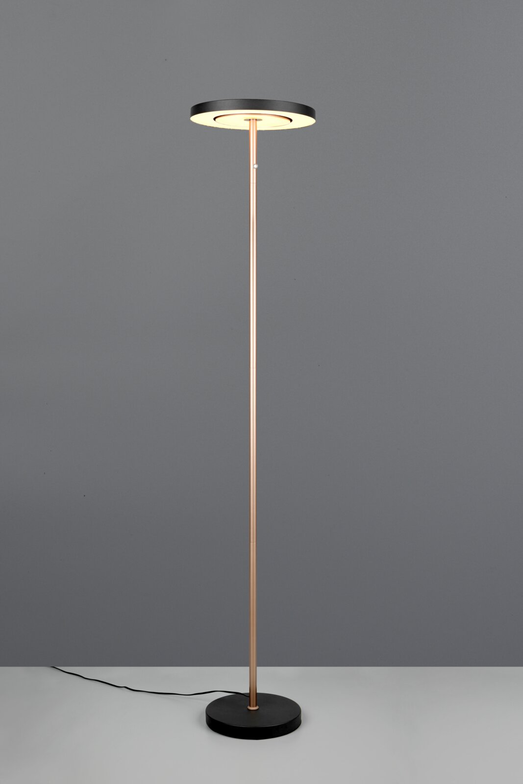 Moderne staande vloerlamp met slanke goudkleurige staander, ronde zwarte voet en schijfvormige lampkop met warm wit licht, frontaal gefotografeerd op grijze achtergrond