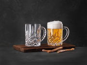 Zwei elegante Bierkrüge aus Kristallglas auf einem Holzbrett, einer gefüllt mit Bier und Schaum, der andere leer, seitliche Perspektive.