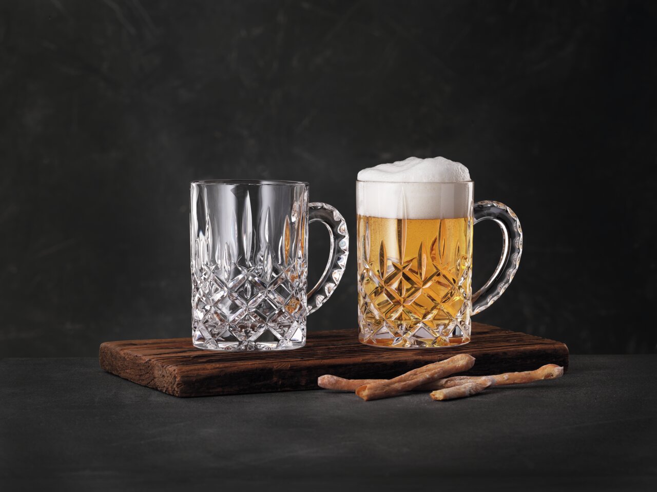 Nachtmann Bierkrug Set NOBLESSE Zwei elegante Bierkrüge aus Kristallglas auf einem Holzbrett, einer gefüllt mit Bier und Schaum, der andere leer, seitliche Perspektive.