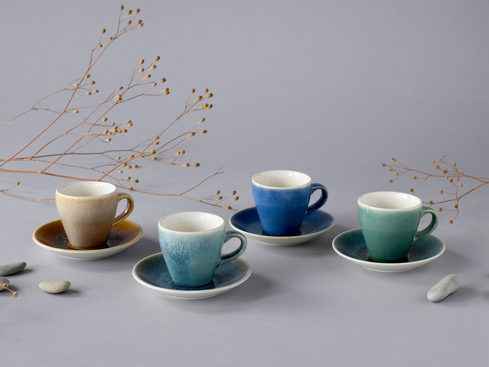 Creatable Espresso-Set 8-tlg CALDERA MIX Vier glasierten Espressotassen mit Untertassen in Blau-, Türkis- und Brauntönen auf grauem Hintergrund, dekoriert mit trockenen Zweigen und Steinen, fotografiert in leicht erhöhter Frontansicht (3/4-Perspektive).