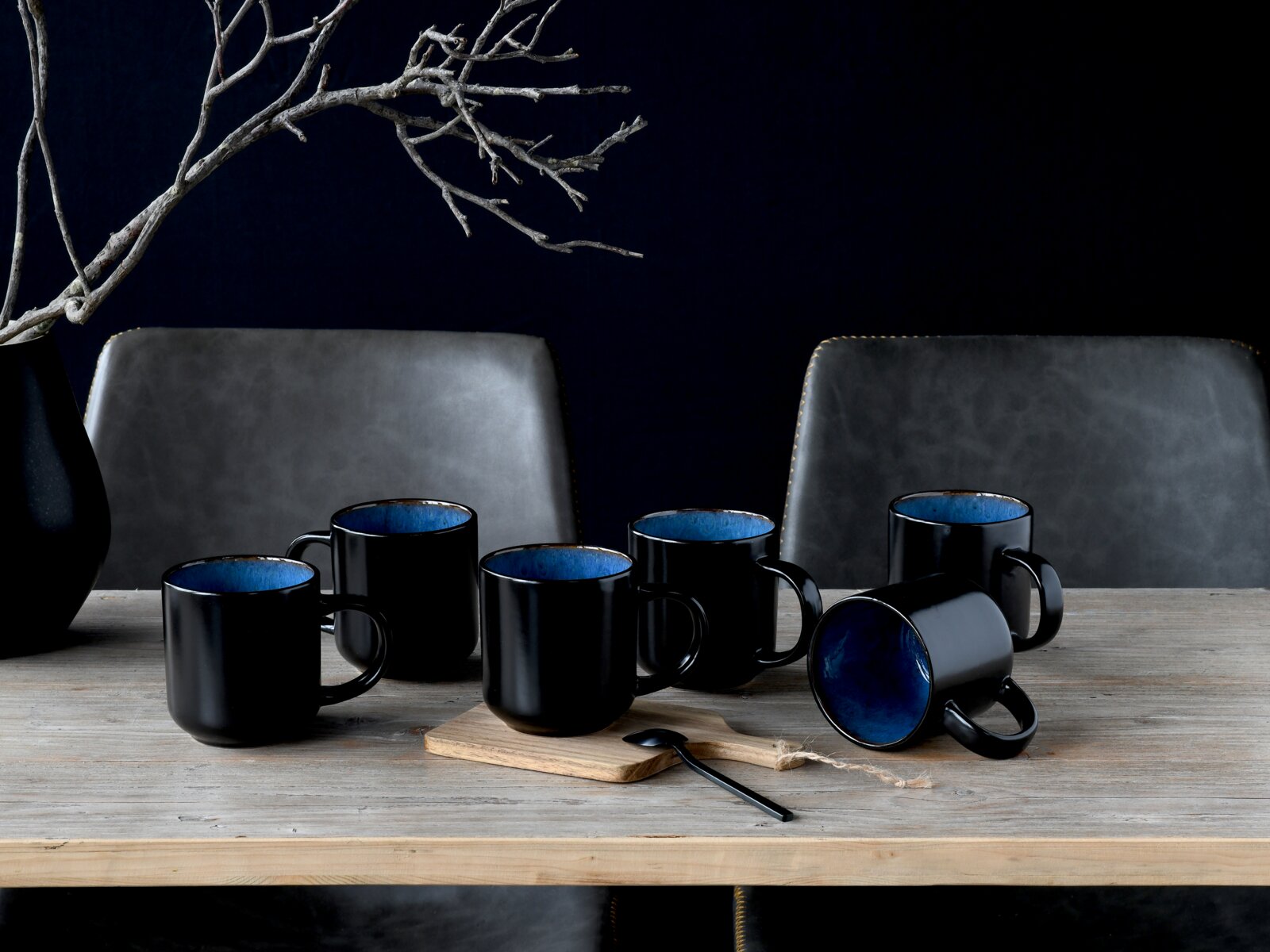 Fünf schwarze Keramik-Kaffeebecher mit blauem Innenraum auf einem Holztisch, ein Becher liegt seitlich, fotografiert in leichter Frontalansicht auf Tischhöhe.