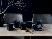 Fünf schwarze Keramik-Kaffeebecher mit blauem Innenraum auf einem Holztisch, ein Becher liegt seitlich, fotografiert in leichter Frontalansicht auf Tischhöhe.