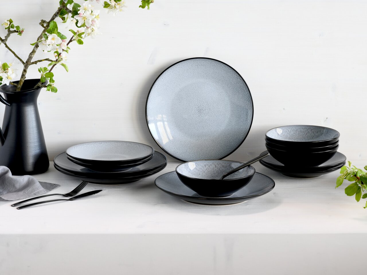 Elegantes 12-teiliges Tellerset in Schwarz und Grau, präsentiert auf einem Tisch mit Blumen im Hintergrund, seitliche Perspektive.