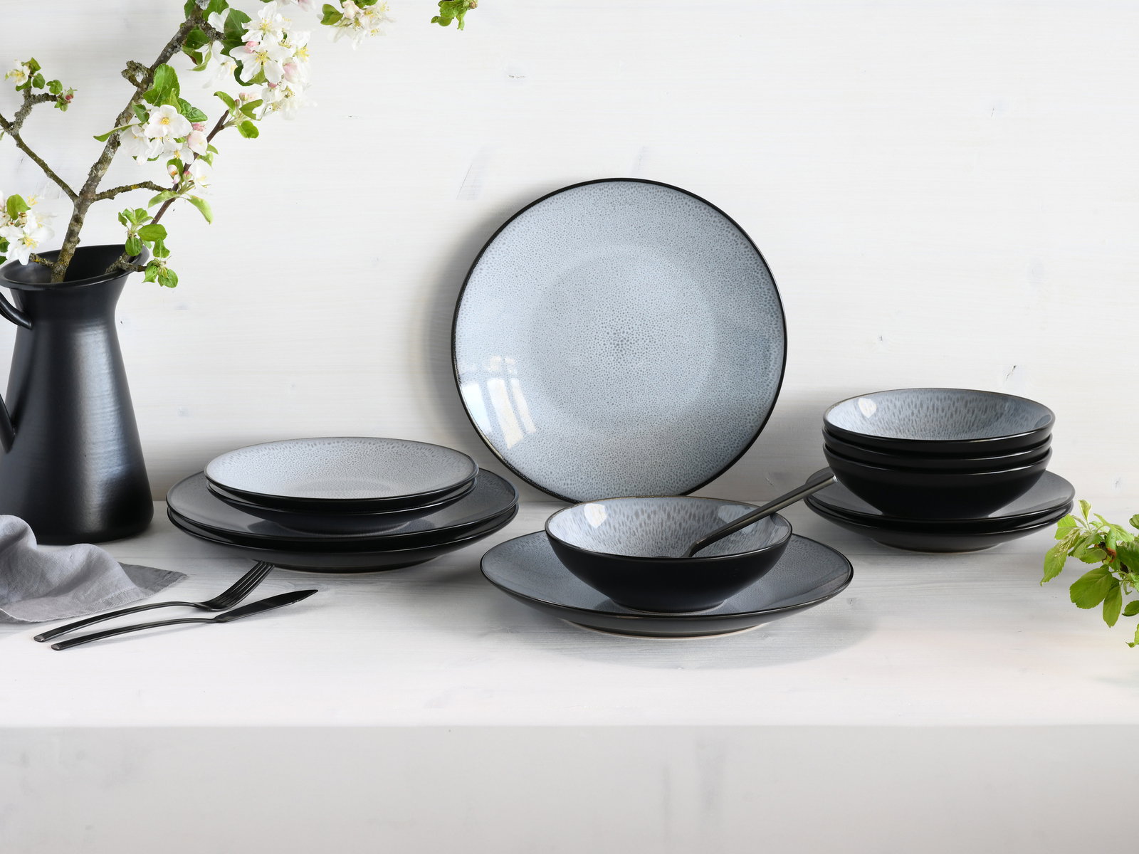 Elegantes 12-teiliges Tellerset in Schwarz und Grau, präsentiert auf einem Tisch mit Blumen im Hintergrund, seitliche Perspektive.