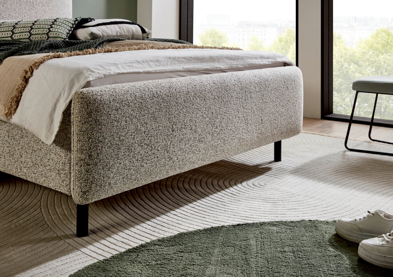 Seitliche Perspektive auf ein modernes Polsterbett in neutralen Farben, umgeben von einem stilvollen Teppich und einem Fenster mit Blick ins Freie.