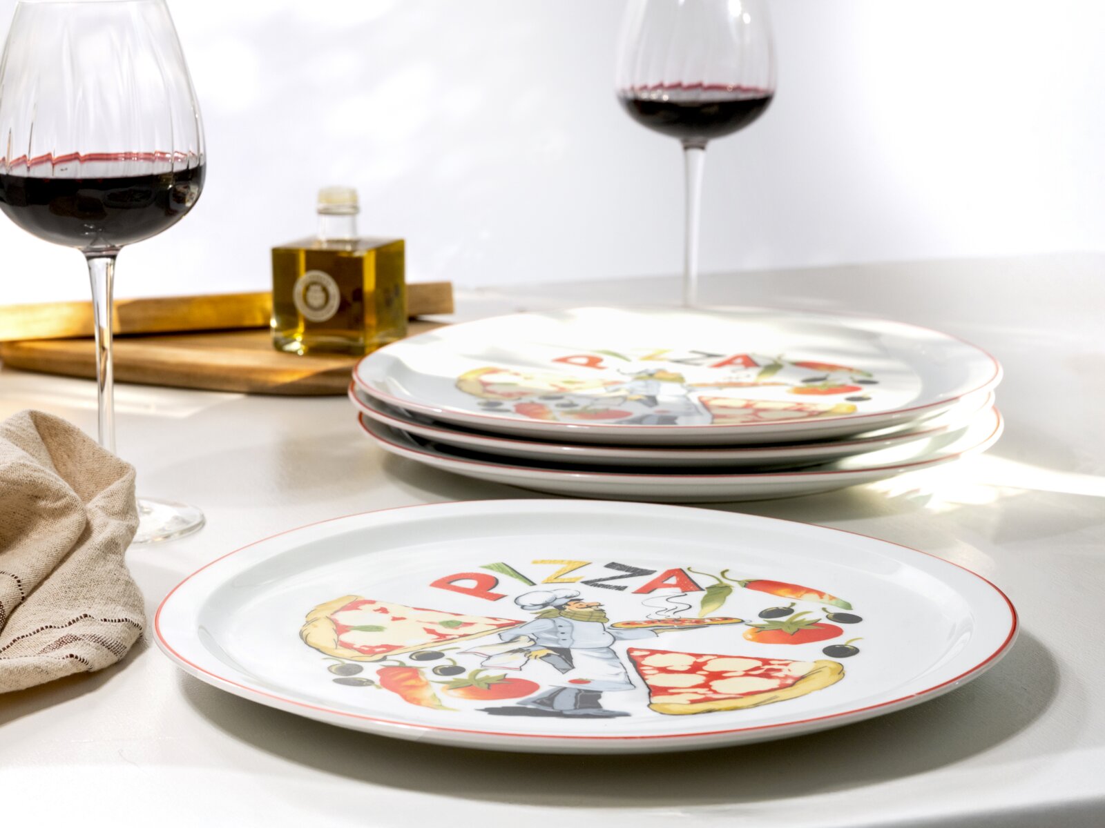 Italienisch dekorierte Pizzateller im Set, gestapelt auf weißem Tisch mit Rotweingläsern und Olivenölflasche im Hintergrund, fotografiert in leicht schräger Seitenansicht auf Tischhöhe (3/4-Perspektive).