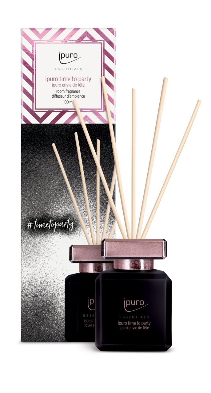 Vorderansicht eines Raumdufts mit schwarzem Flakon und rosa Deckel, umgeben von Duftstäbchen, vor einer dekorativen Verpackung mit pink-weißen Streifen.