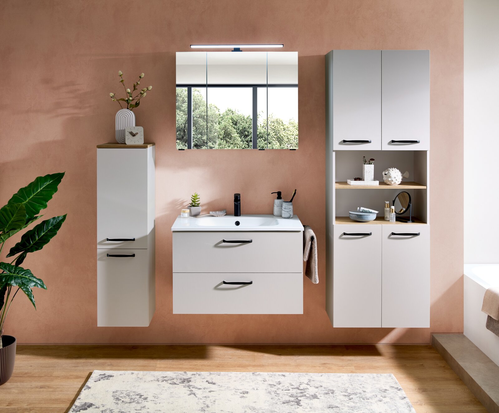 Frontale Ansicht eines modernen, wandhängenden Badmöbel-Sets in Weiß mit Waschtisch, Spiegelschrank mit LED-Leuchte sowie Hochschrank und Regal-/Schrankkombination, schwarze Griffe vor roséfarbener Wand