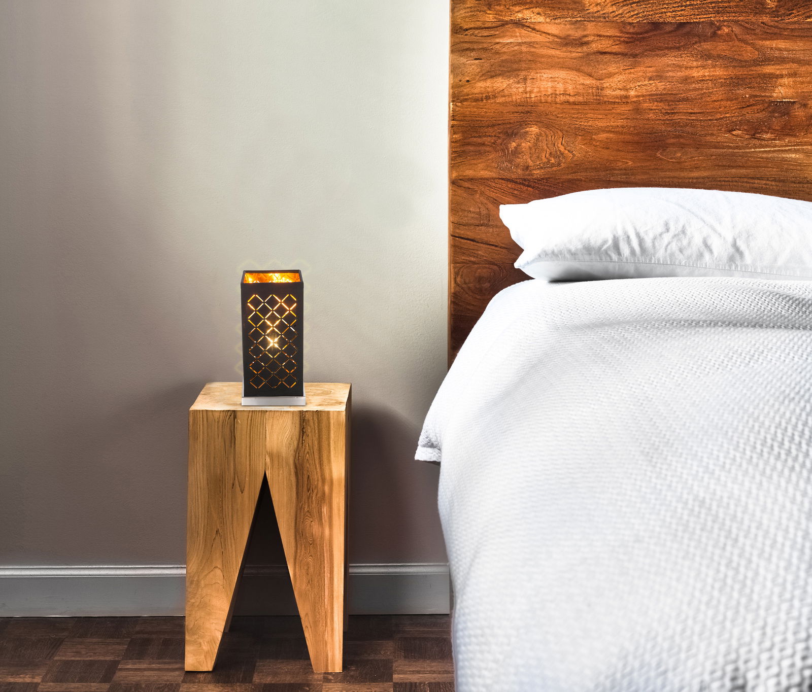 Moderne Tischleuchte mit geometrischem Muster auf einem Holztisch neben einem Bett, seitliche Perspektive.