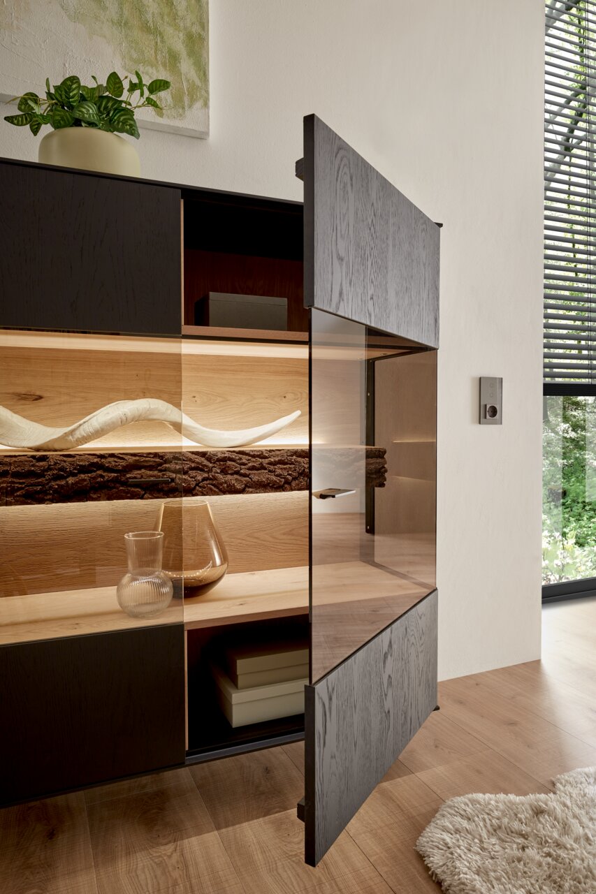 Geöffnetes Highboard mit dunkler Holzfront, dekorativen Elementen und Pflanzen, seitliche Perspektive