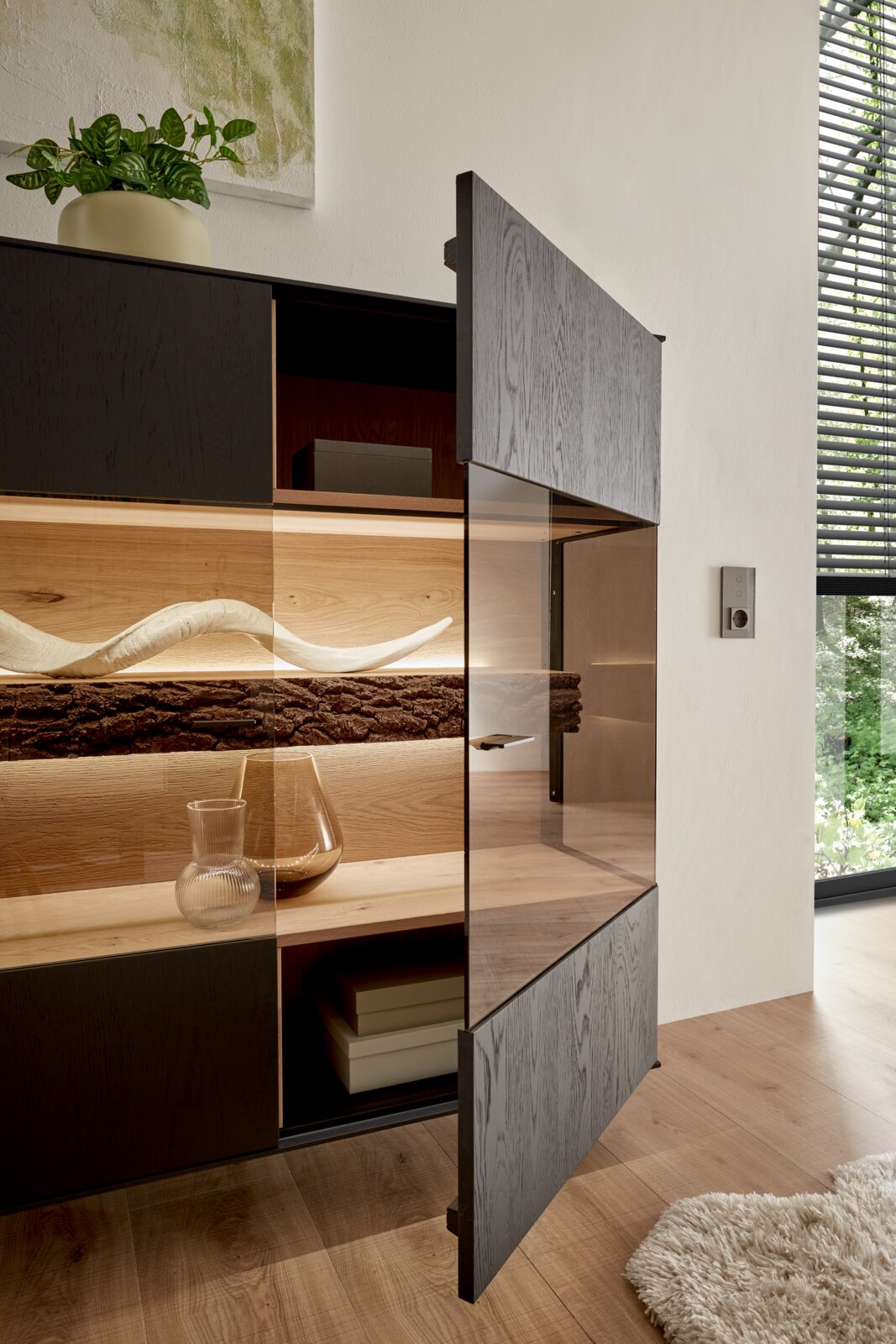Geöffnetes Highboard mit dunkler Holzfront, dekorativen Elementen und Pflanzen, seitliche Perspektive