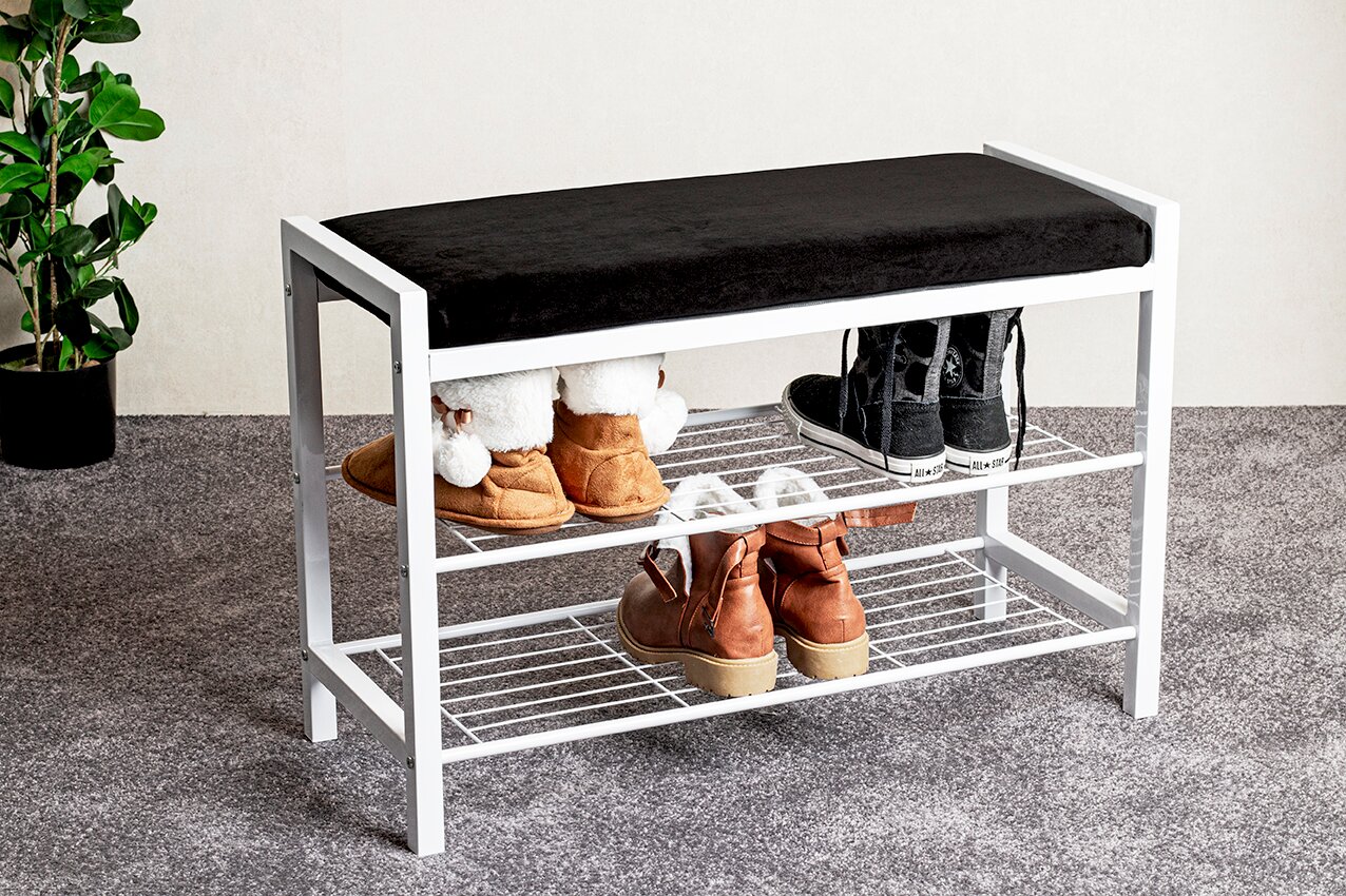 Weiße Schuhbank mit schwarzem Sitzpolster und zwei Drahtablagen, bestückt mit Schuhen, im Eingangsbereich auf grauem Teppich – Perspektive: seitliche Frontansicht leicht von oben.