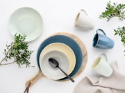 Draufsicht (Flatlay) auf modernes Keramikgeschirr in Blau-, Creme- und Minttönen: gestapelte Teller und Schalen auf Holzbrett mit schwarzem Löffel, dazu drei Tassen, Leinentuch und grüne Zweige auf weißem Hintergrund.