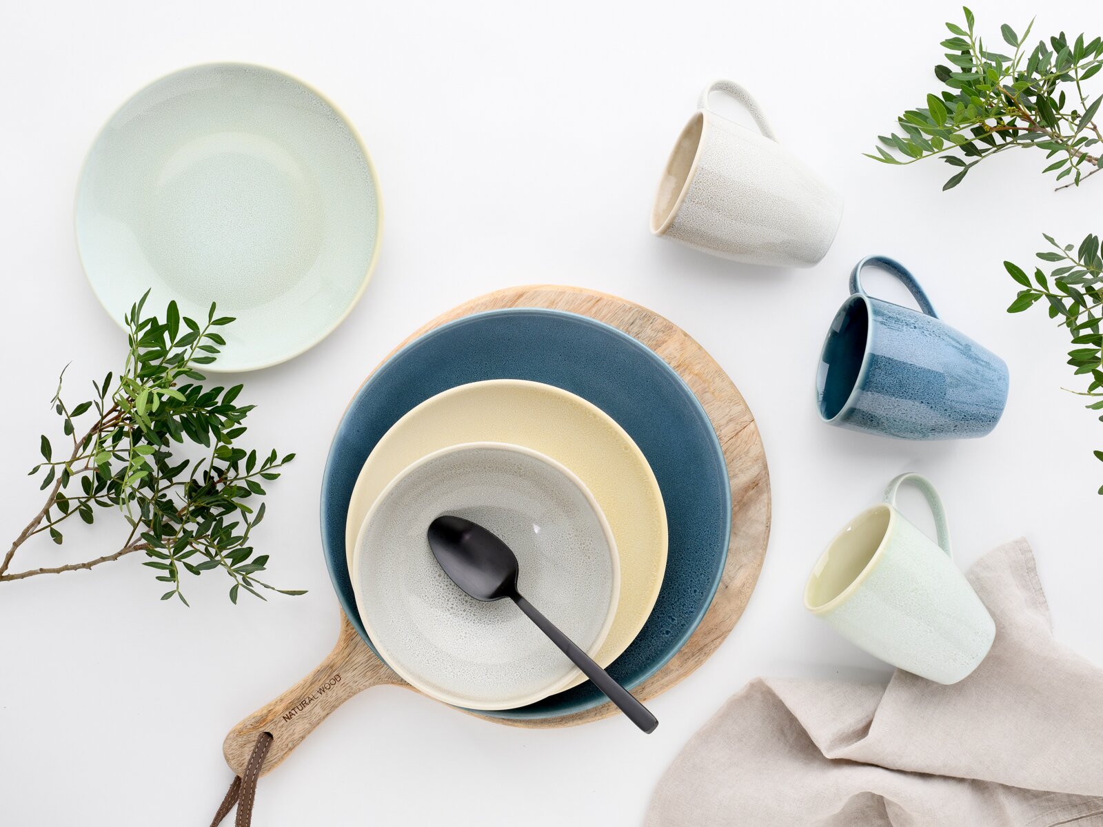 Draufsicht (Flatlay) auf modernes Keramikgeschirr in Blau-, Creme- und Minttönen: gestapelte Teller und Schalen auf Holzbrett mit schwarzem Löffel, dazu drei Tassen, Leinentuch und grüne Zweige auf weißem Hintergrund.