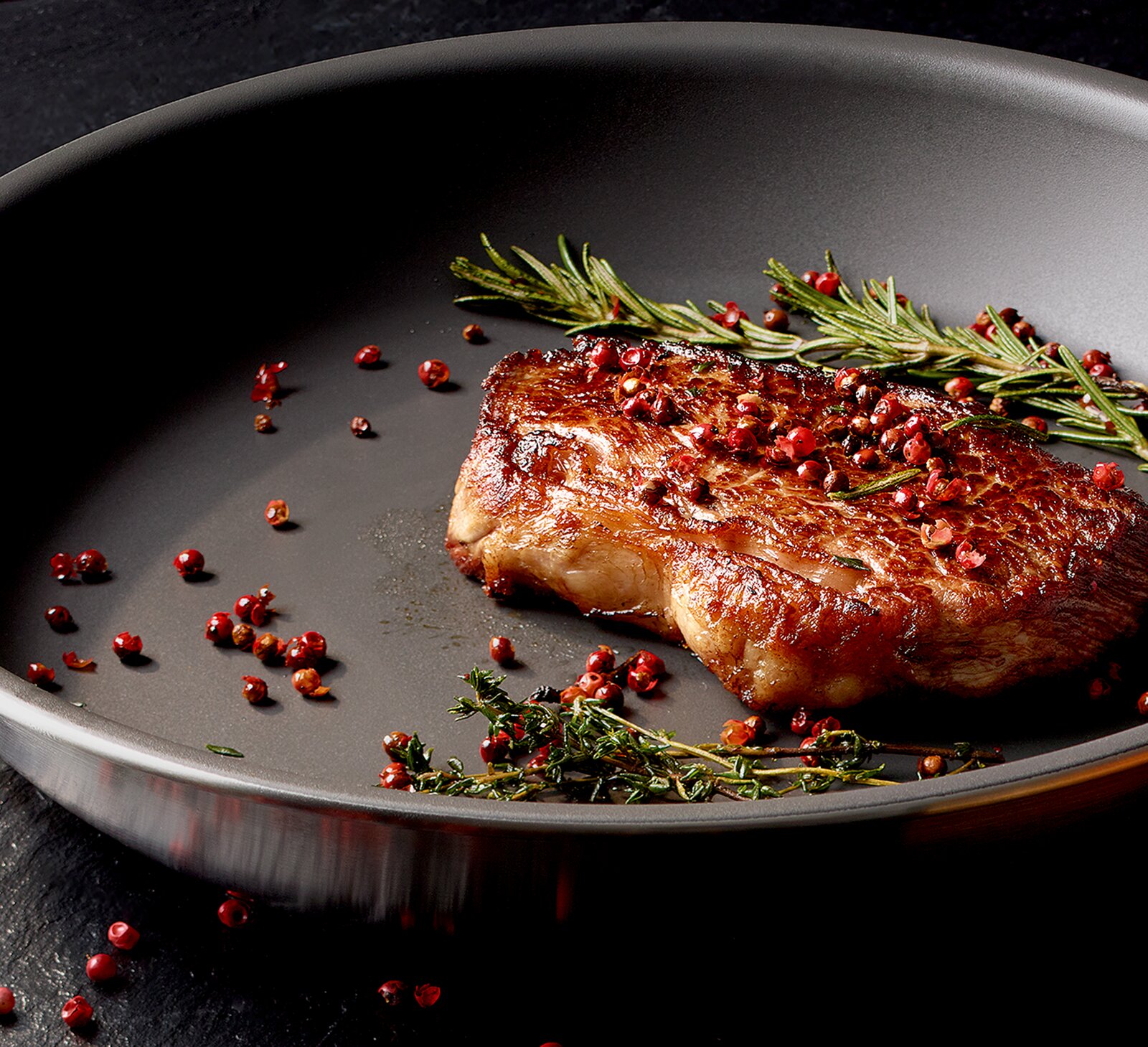 Close-up vanuit een licht schuin bovenaanzicht van een gebakken steak in een zwarte koekenpan, gegarneerd met rozemarijn, tijm en roze peperkorrels.
