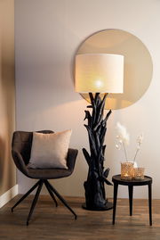 Stehlampe mit natürlichem Holzfuß und hellem Lampenschirm, seitliche Perspektive, neben einem Sessel und einem kleinen Tisch mit Dekoration.