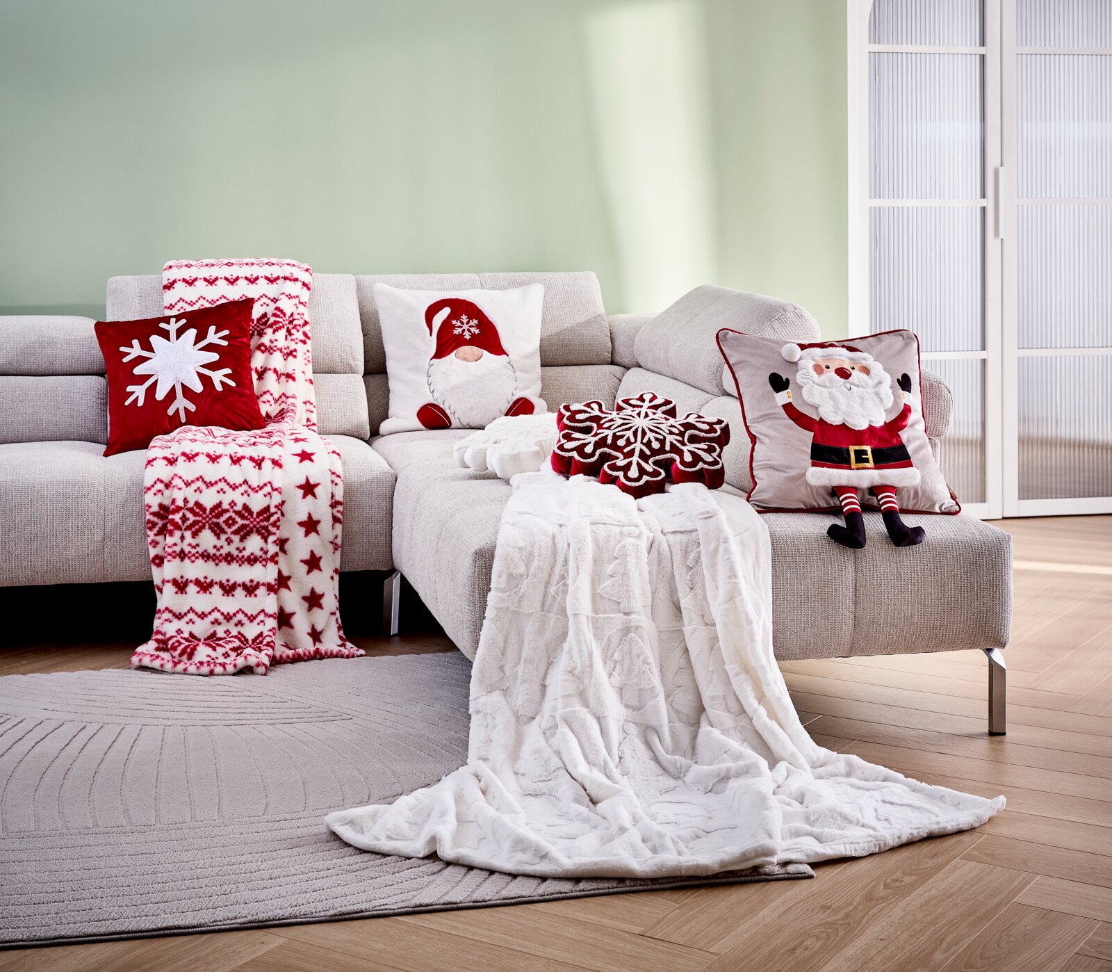 Een gezellige woonkamerscène met een witte knuffeldeken op een lichte bank, omringd door kussens in rood en wit die in kerstsfeer zijn versierd. Aanzicht van voren.