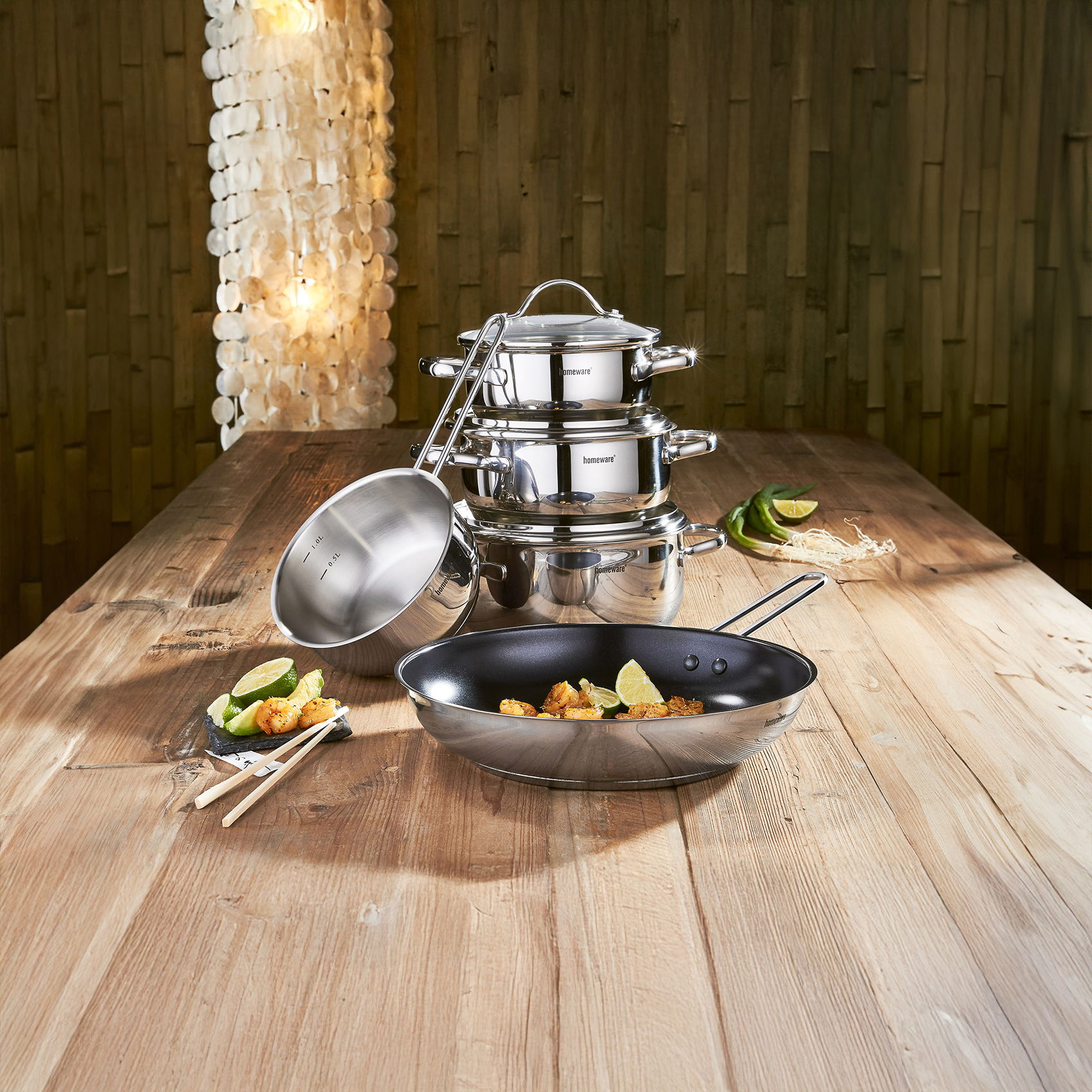 Edelstahl-Topfset und Pfanne auf Holztisch, dekoriert mit Speisen, Limetten und Gemüse in warm beleuchteter Küche