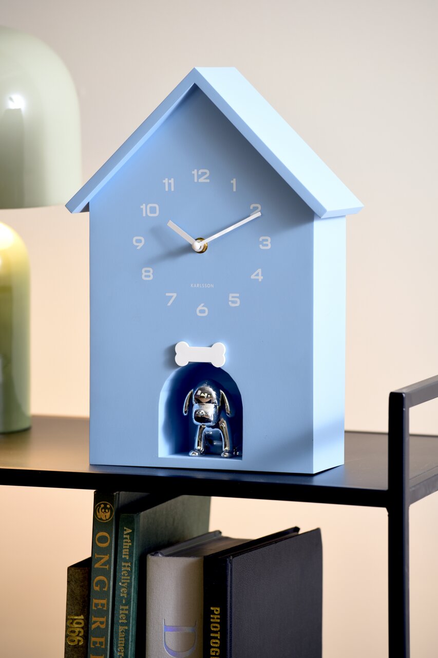 Blaue Wanduhr in Form eines Hauses mit einem silbernen Hund in einer kleinen Nische und einem weißen Knochen darüber, seitliche Perspektive.