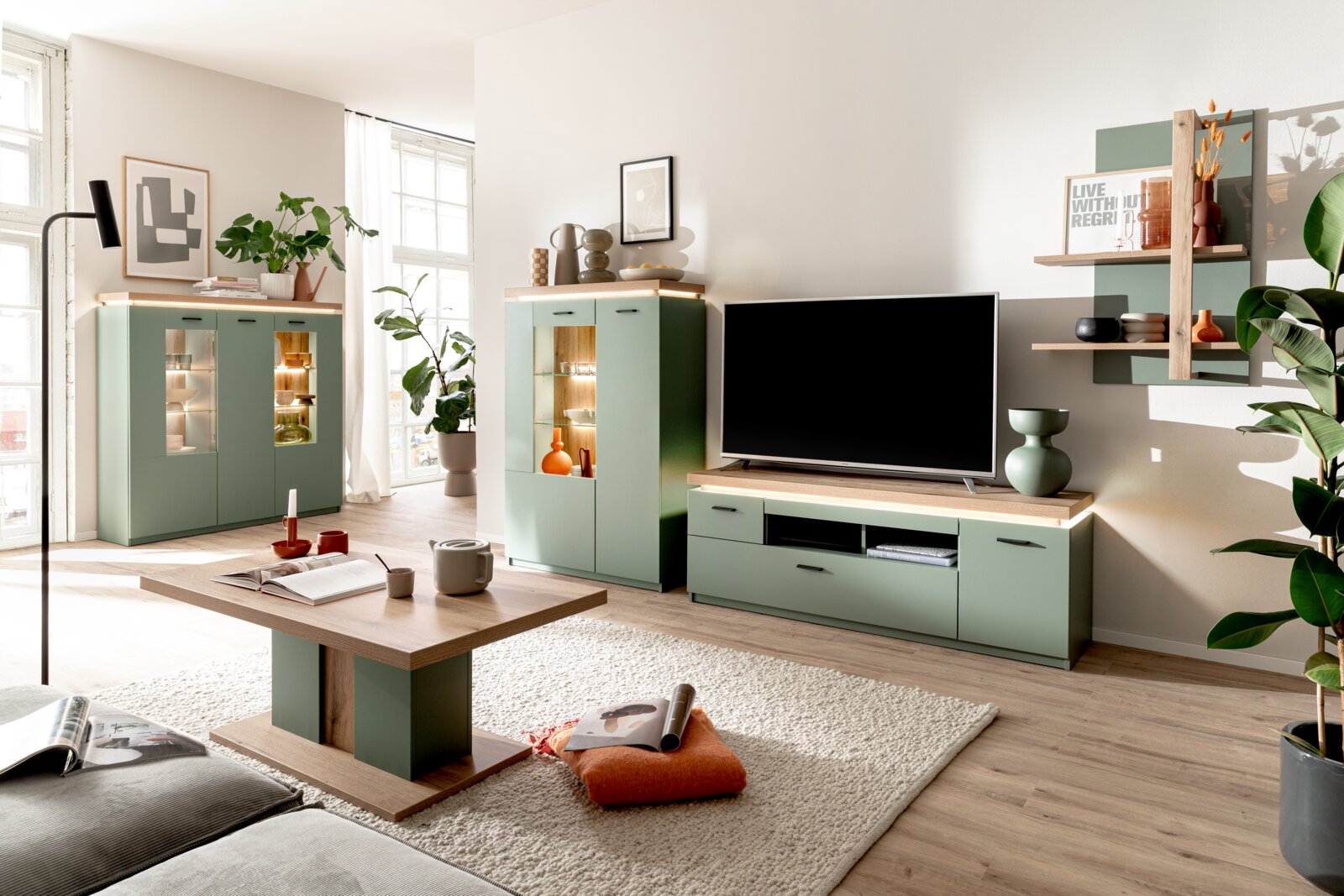 Z2 TV-Lowboard MONITZ Modernes Wohnzimmer-Set in Salbeigrün und Holzoptik mit TV-Lowboard, zwei Vitrinenschränken mit Beleuchtung, Couchtisch und Wandregal; Ansicht aus leicht erhöhter Frontperspektive im hellen Raum