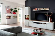 Modernes Wohnzimmer-Set mit weißem TV-Lowboard, Hängeregal, Vitrine und Sideboard mit Holzoptik-Akzenten vor dunkler Akzentwand, perspektivisch in schräger Frontansicht aufgenommen