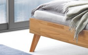 Detailfoto van een modern bedframe van massief hout in een lichte houtlook met schuine houten poten, een wit matras en een grijze deken; perspectief: close-up van opzij, van onderaf.