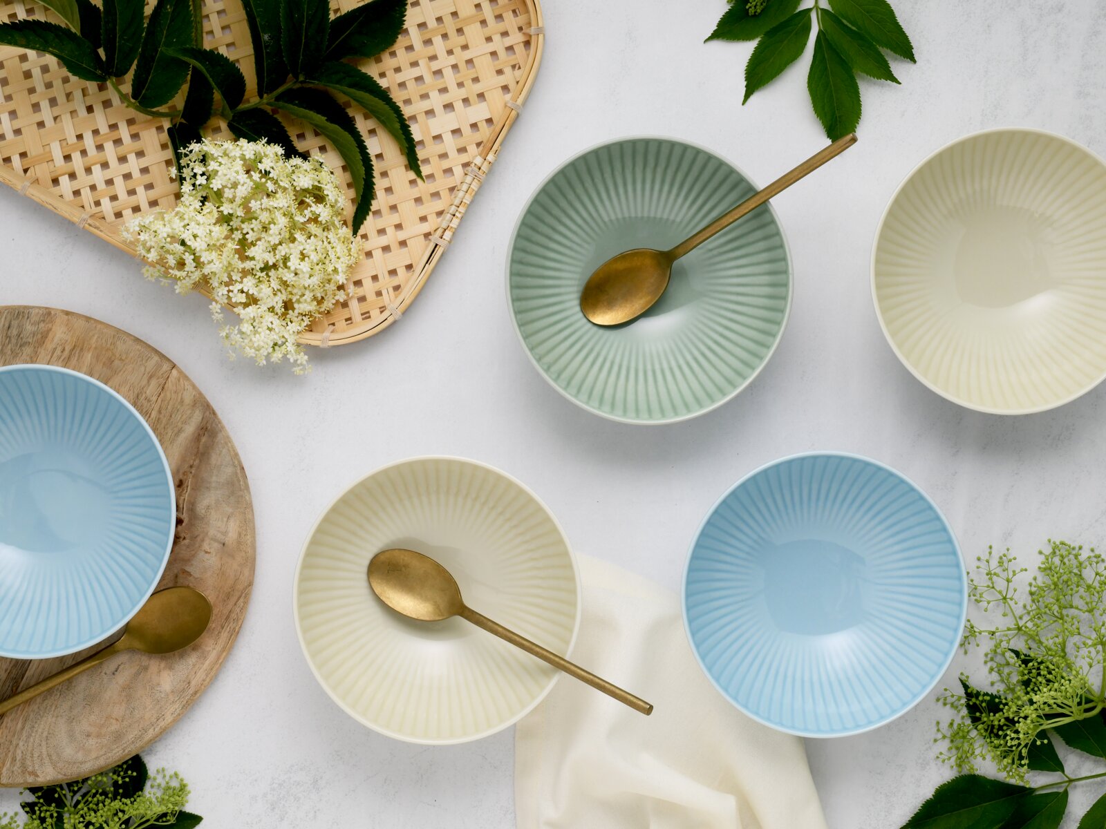 Draufsicht auf pastellfarbene, geriffelte Keramikschalen in Blau, Mint und Creme mit goldfarbenen Löffeln, dekoriert mit Blumen und Blättern auf hellem Hintergrund
