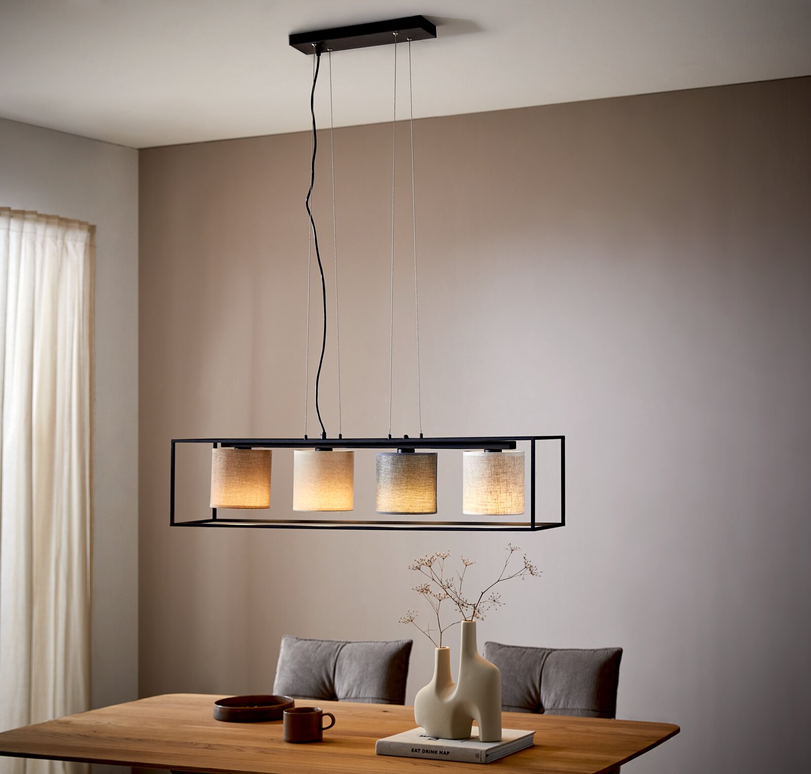 Moderne Hängeleuchte mit vier zylindrischen Lampenschirmen in einem rechteckigen Rahmen, von der Decke hängend über einem Esstisch, seitliche Perspektive.