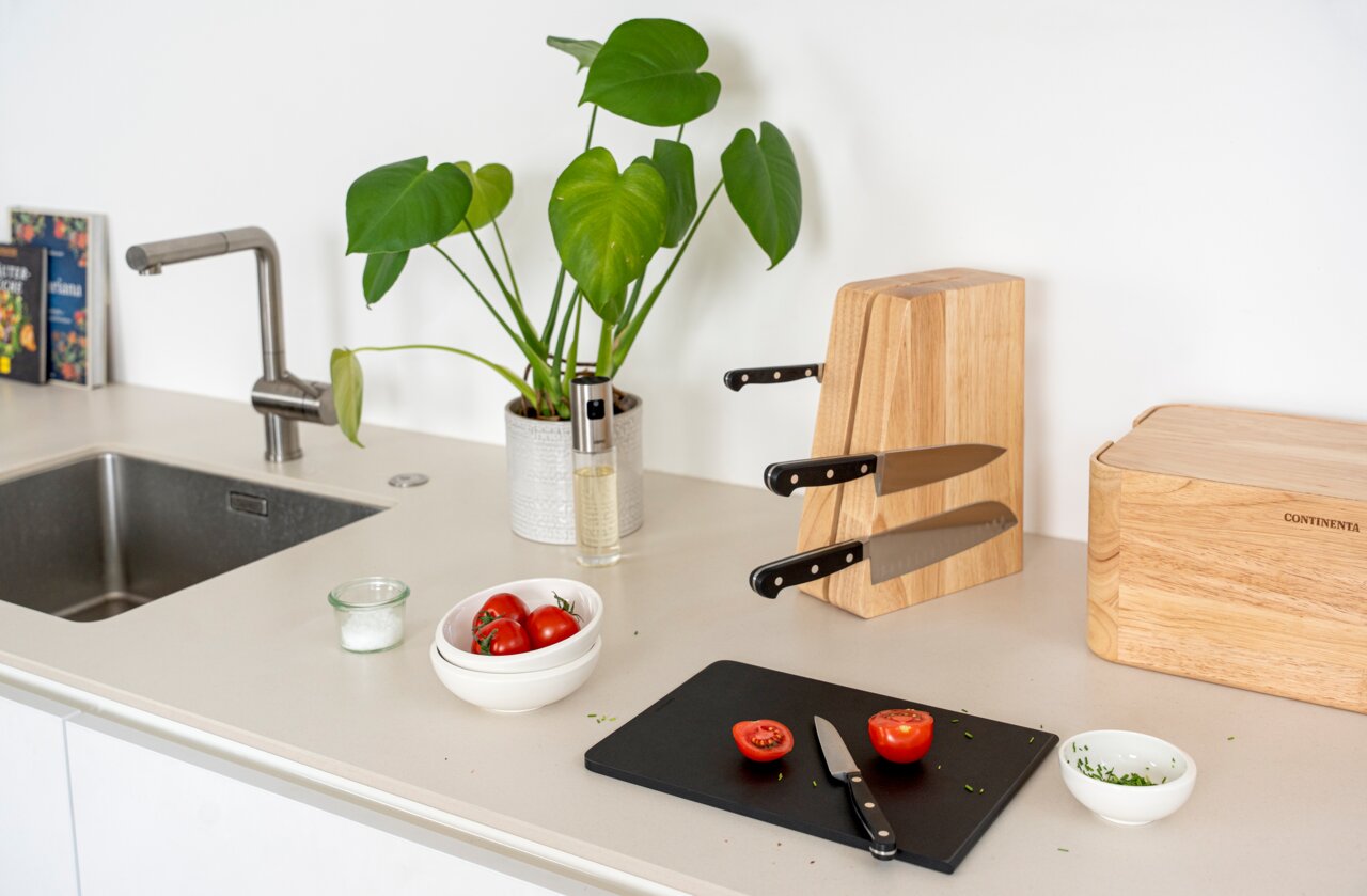 Continenta Magnetisch messenblok Modern keukenblad in schuin vooraanzicht met een houten messenblok en drie messen, een zwarte snijplank met gehalveerde tomaten en een mes, een schaal met tomaten, kruiden en zout, en een kamerplant naast de gootsteen.