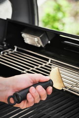 Hand hält einen Universalschaber ScrapX mit schwarzem Griff und goldener Klinge, der auf einem Grillrost verwendet wird. Perspektive von der Seite.