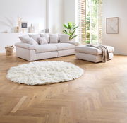 Großes, helles Big-Sofa mit mehreren Kissen in einem modernen Wohnzimmer, seitliche Perspektive. Daneben ein passender Hocker und ein flauschiger, runder Teppich auf Holzboden.