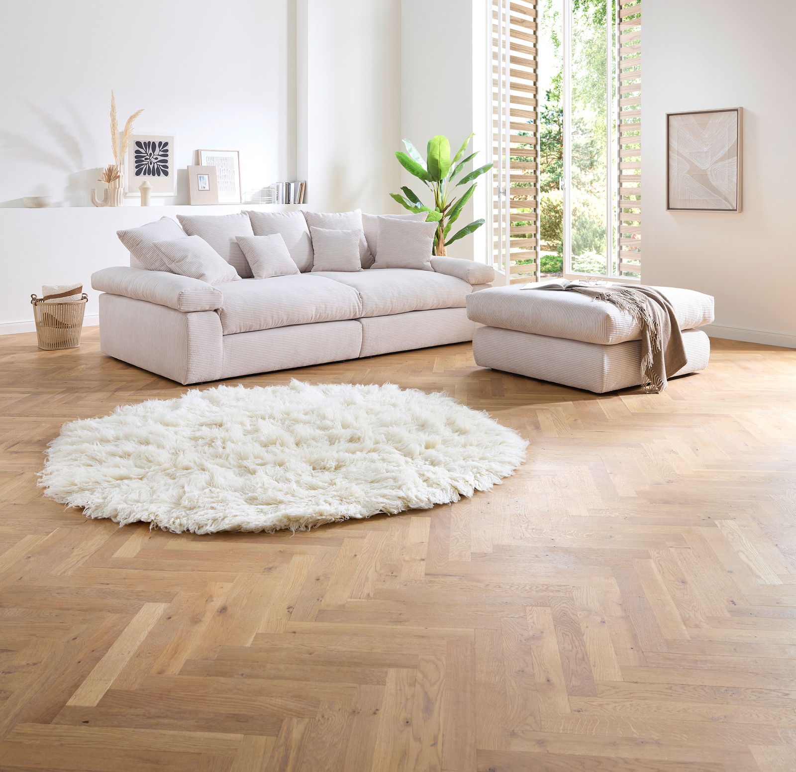 Großes, helles Big-Sofa mit mehreren Kissen in einem modernen Wohnzimmer, seitliche Perspektive. Daneben ein passender Hocker und ein flauschiger, runder Teppich auf Holzboden.