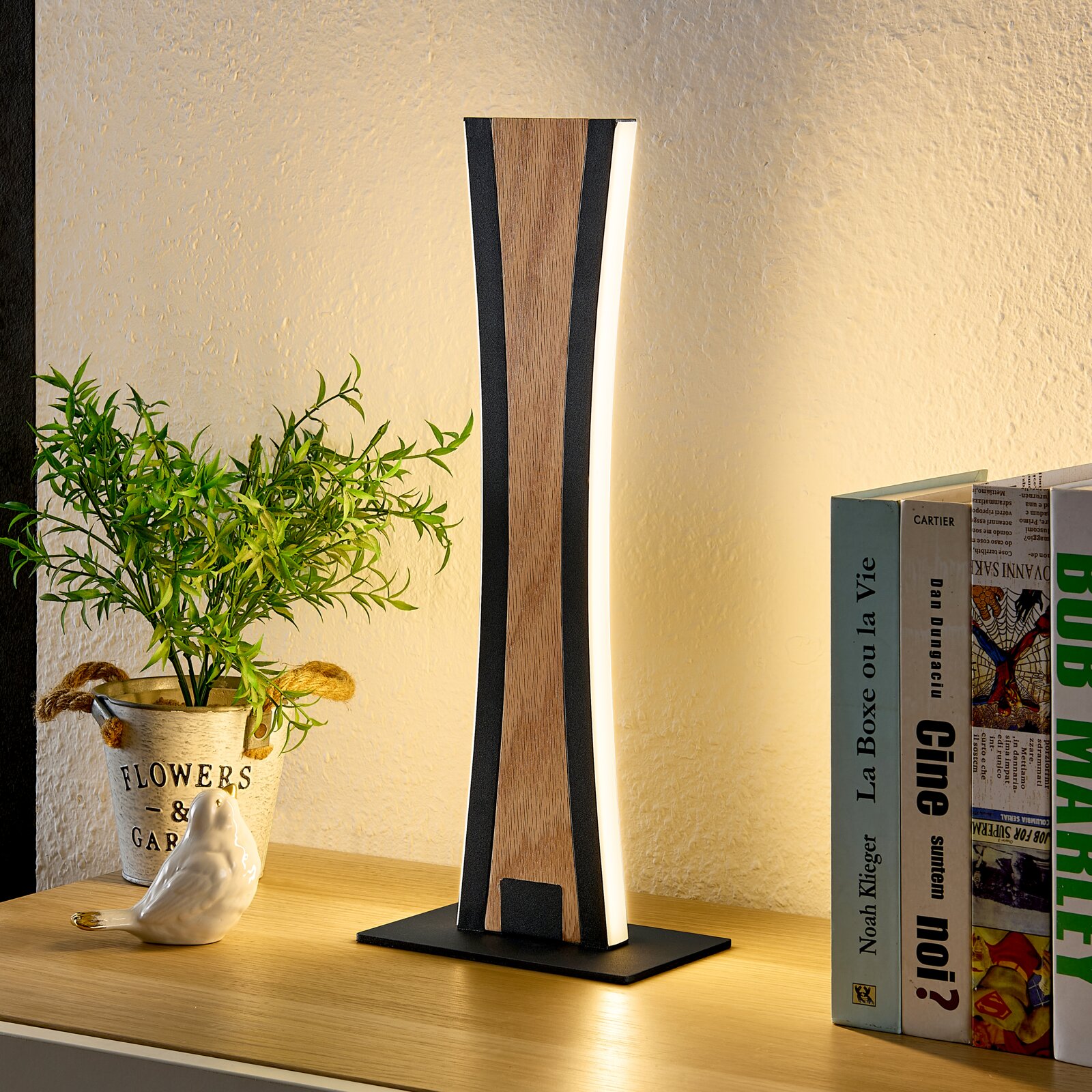 Moderne LED-Tischleuchte mit Holzoptik und schwarzem Rahmen auf einem Sideboard, warmweiß leuchtend, perspektivisch frontal leicht seitlich aufgenommen; daneben Bücher und Pflanze