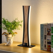 Moderne LED-Tischleuchte mit Holzoptik und schwarzem Rahmen auf einem Sideboard, warmweiß leuchtend, perspektivisch frontal leicht seitlich aufgenommen; daneben Bücher und Pflanze
