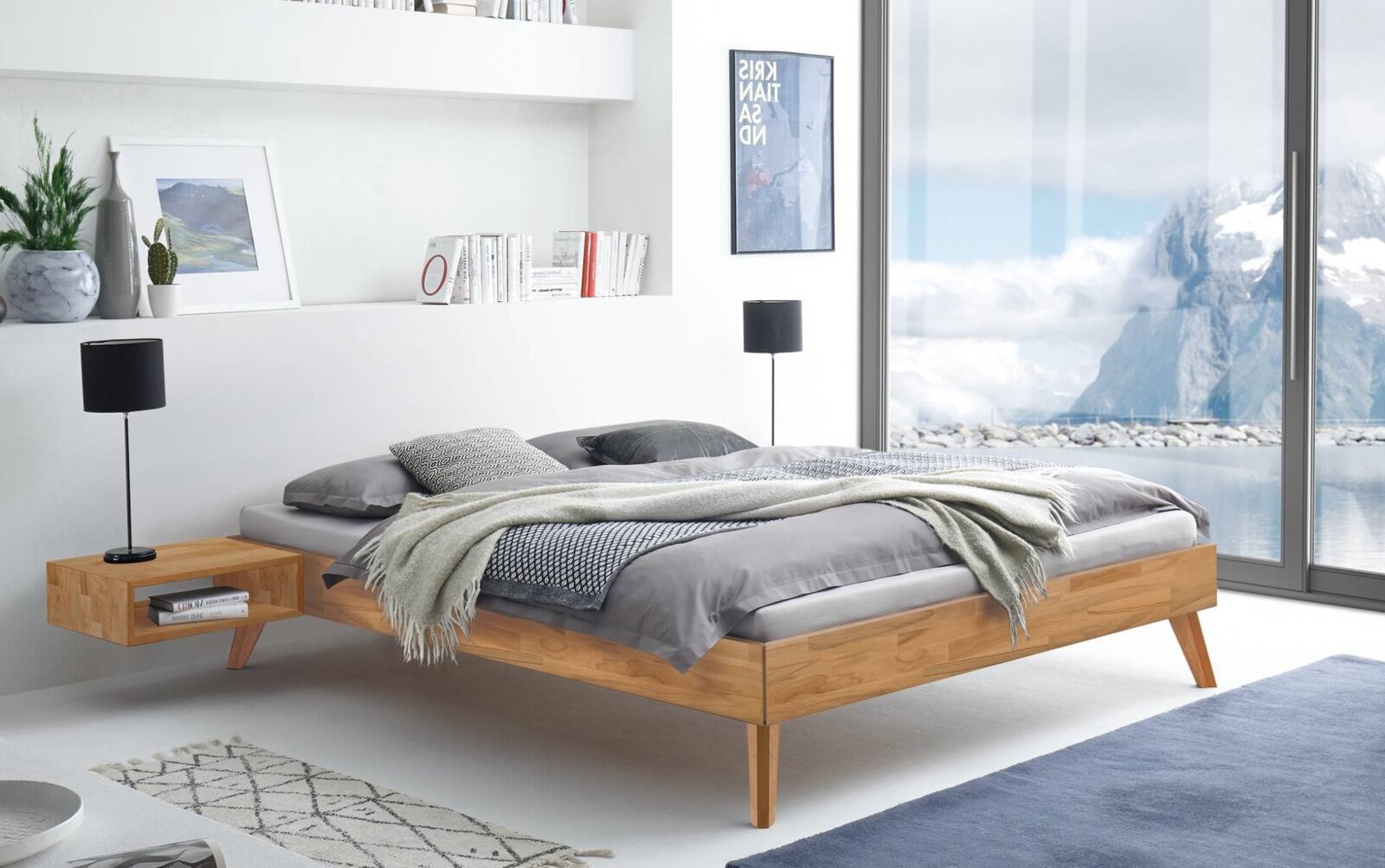 Modern houten bed in Scandinavische stijl met een laag frame en schuine poten, in een lichte slaapkamer met grijs beddengoed – perspectief: zijaanzicht, licht van links.
