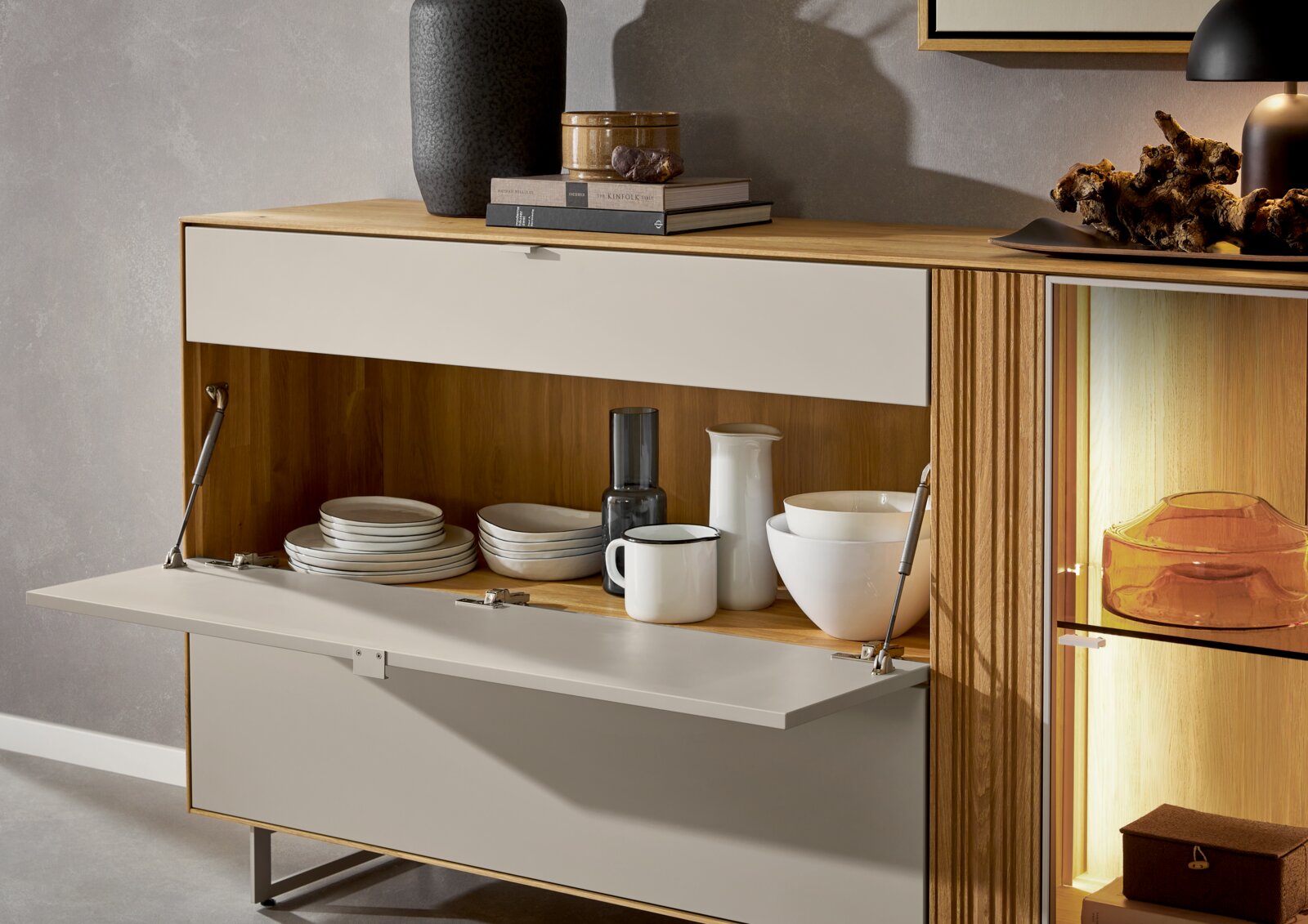 Modernes Sideboard aus Holz mit hellgrauen Klappen, offene Klappe zeigt Geschirr im Fach; rechts beleuchtete Vitrine mit Glasablage, Ansicht schräg von vorne.