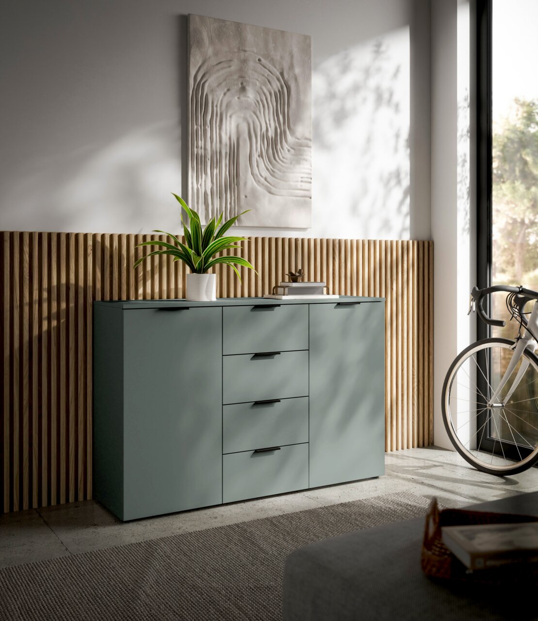 Modernes Sideboard in mattem Grün mit vier Schubladen und zwei Türen, dekoriert mit einer Pflanze und Kunstwerk an der Wand, seitliche Perspektive.
