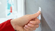 Nahaufnahme einer Hand, die einen tesa Powerstrip Waterproof an einer glatten, grauen Wand befestigt.