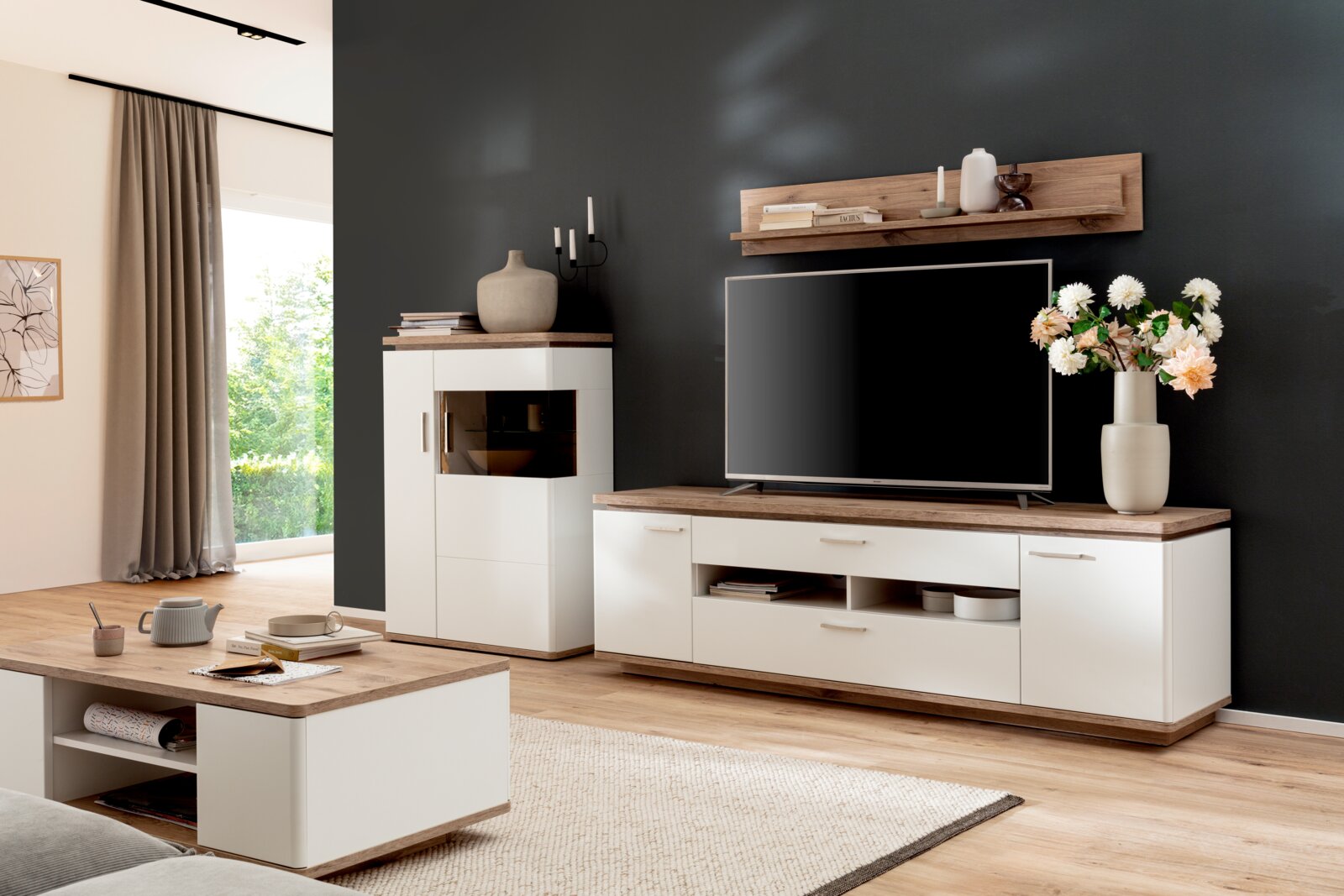 Moderne woonkamerset in wit en eikenlook, bestaande uit een tv-kast, vitrinekast, salontafel en wandplank, geplaatst voor een donkere muur, in perspectief vanuit een licht zijdelings vooraanzicht