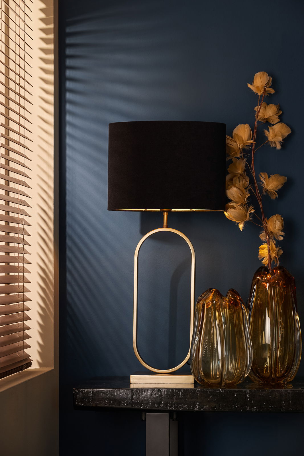 Eleganter Tisch mit einem ovalen Lampenschirm in Schwarz und goldener Basis, daneben dekorative Glasvasen und gelbe Trockenblumen, seitliche Perspektive.