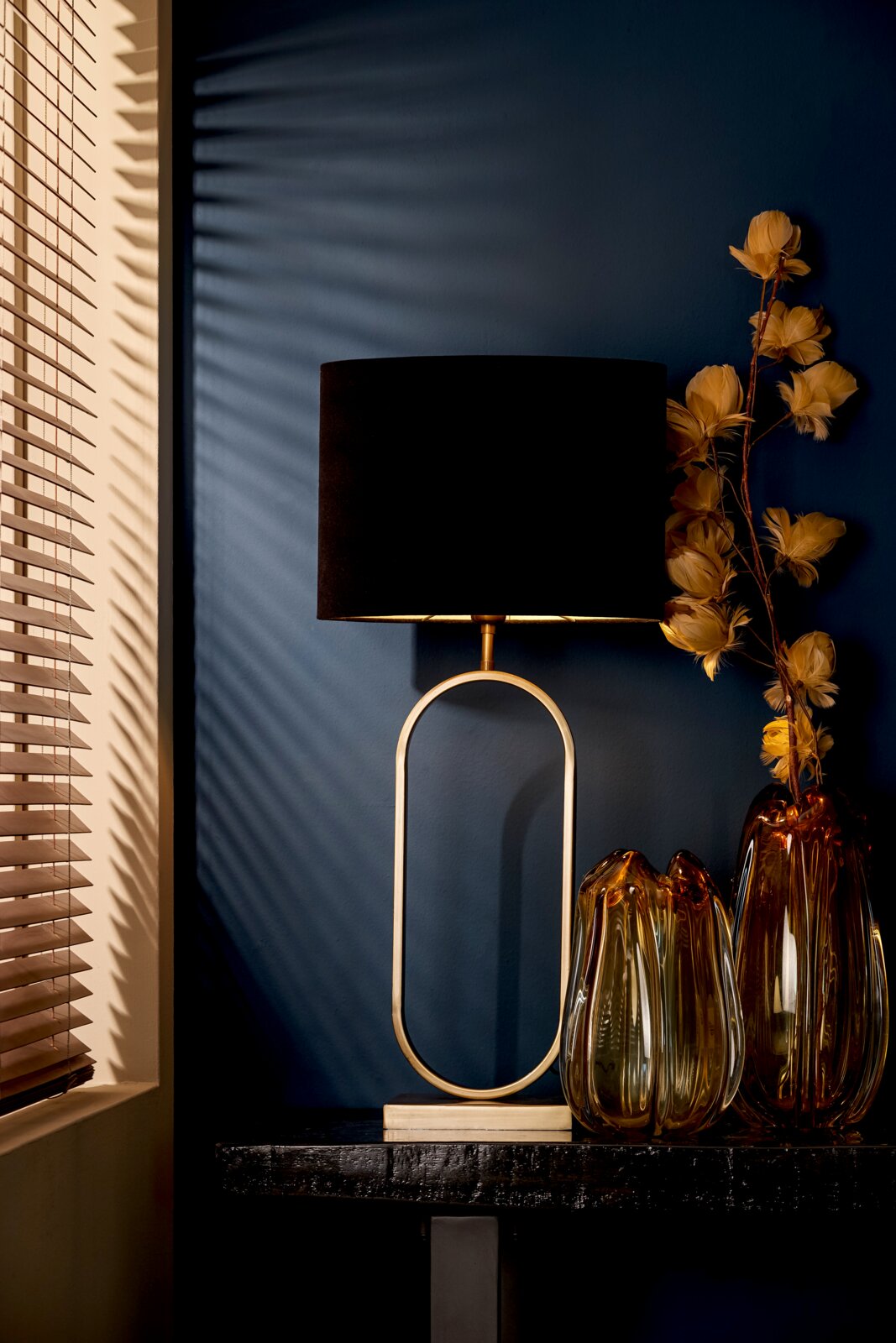Eleganter Tisch mit einem ovalen Lampenschirm in Schwarz und goldener Basis, daneben dekorative Glasvasen und gelbe Trockenblumen, seitliche Perspektive.