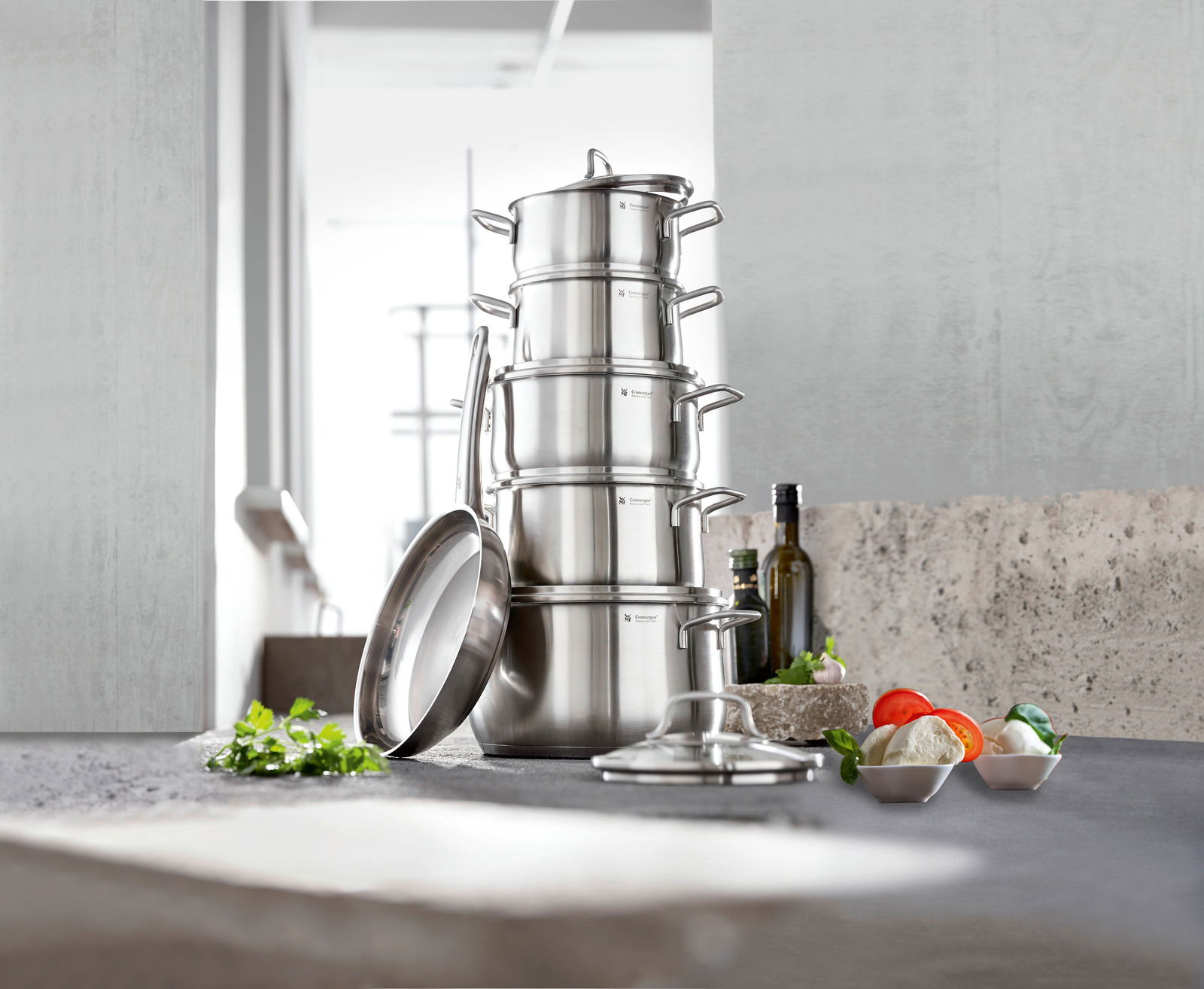 Stapel von Edelstahlkochtöpfen mit Deckeln, seitliche Perspektive, in einer modernen Küche mit frischen Zutaten im Vordergrund.