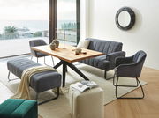 Wohnzimmer mit modernem Esstisch aus Holz, umgeben von grauen Polstermöbeln, darunter ein Armlehnstuhl, eine Sitzbank und ein Sofa. Im Hintergrund ist ein großer Spiegel an der Wand und ein Blick auf das Meer durch die Fenster. Perspektive: seitlich von v