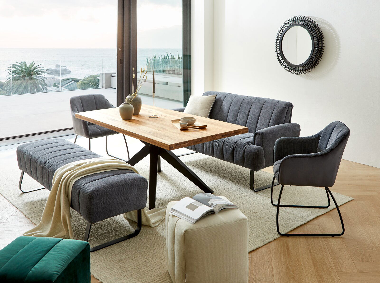 Novel Hocker LAILA Wohnzimmer mit modernem Esstisch aus Holz, umgeben von grauen Polstermöbeln, darunter ein Armlehnstuhl, eine Sitzbank und ein Sofa. Im Hintergrund ist ein großer Spiegel an der Wand und ein Blick auf das Meer durch die Fenster. Perspektive: seitlich von v