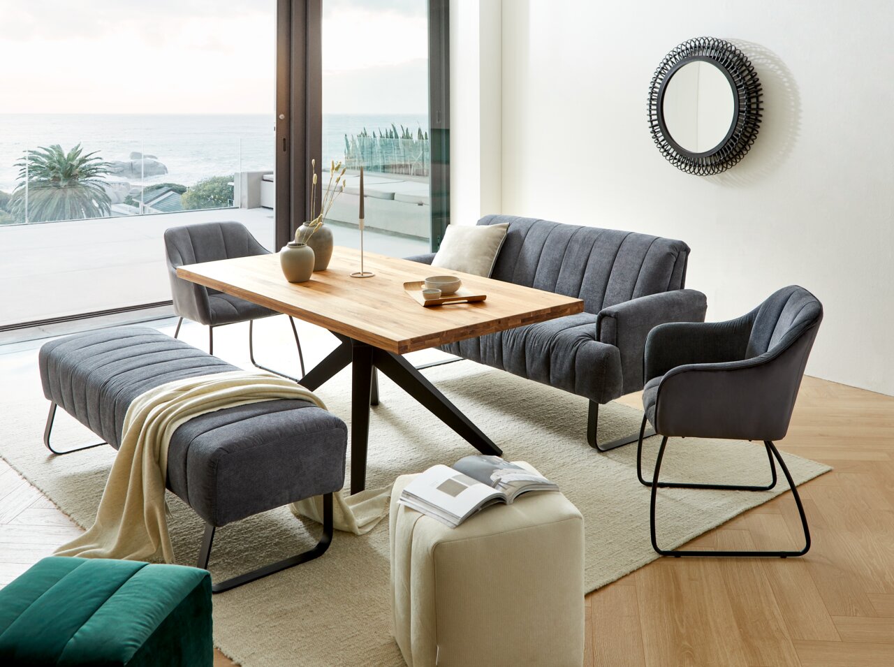 Wohnzimmer mit modernem Esstisch aus Holz, umgeben von grauen Polstermöbeln, darunter ein Armlehnstuhl, eine Sitzbank und ein Sofa. Im Hintergrund ist ein großer Spiegel an der Wand und ein Blick auf das Meer durch die Fenster. Perspektive: seitlich von v
